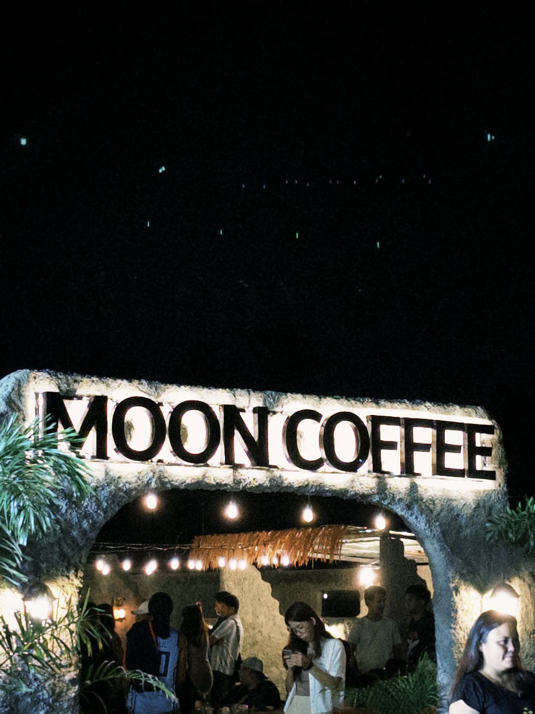 The iconic Moon Coffee stone arch entrance lit at night, crowd gathered below the illuminated sign