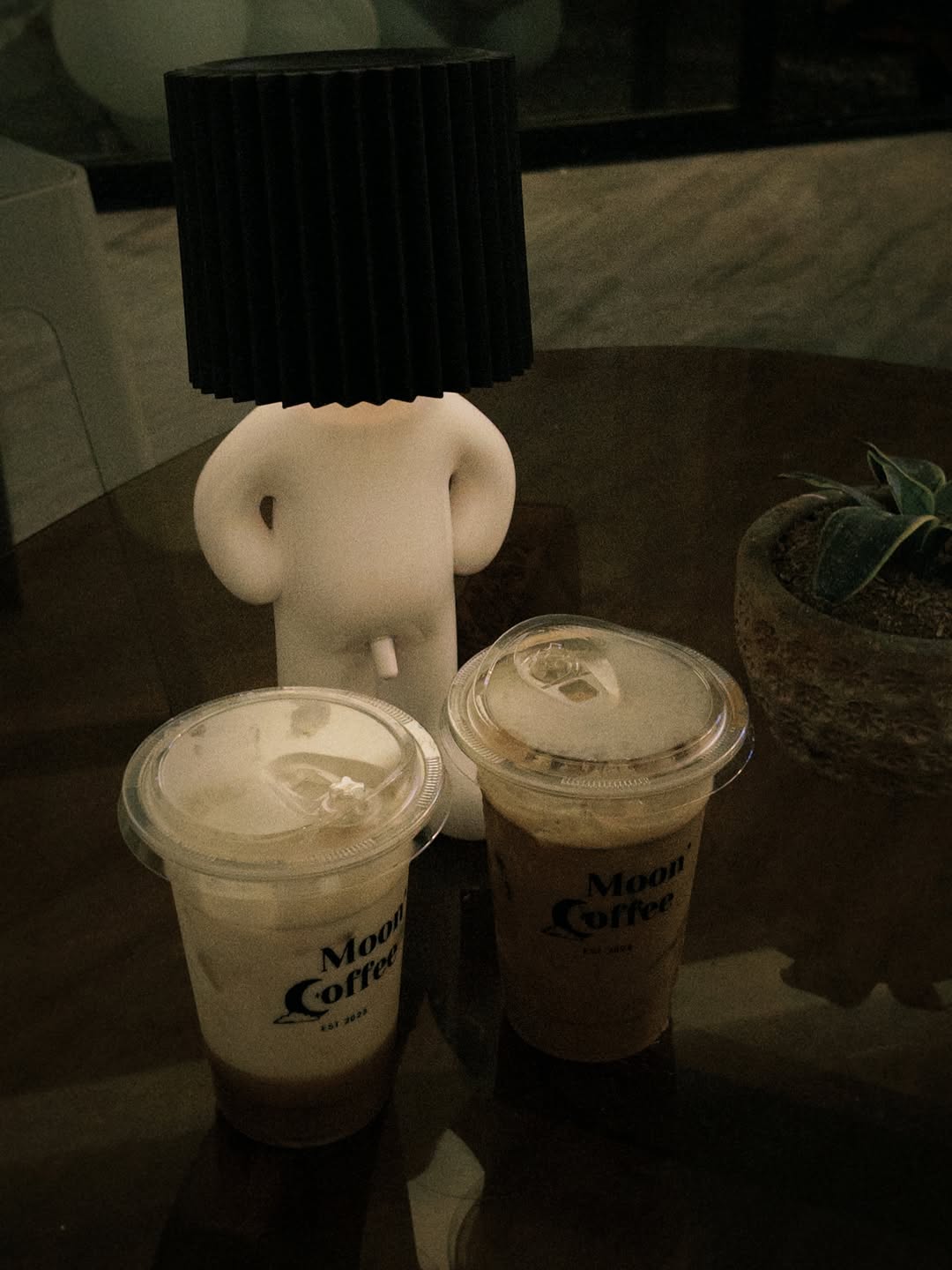 Two Moon Coffee branded cups beside the signature lamp figurine on a dark table — moody interior atmosphere