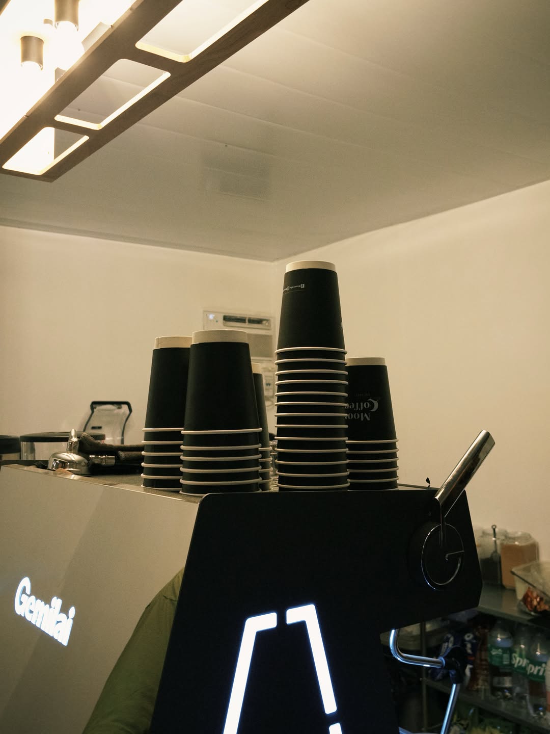 Inside the Moon Coffee 2.0 bar — a Gemilai espresso machine with stacks of branded black paper cups and the barista at work