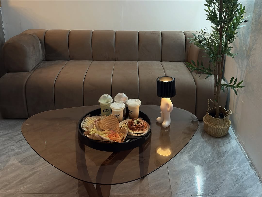 Moon Coffee 2.0 indoor seating area — velvet sofa, glass table with food tray and branded cups, olive tree in the corner